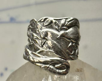 Puede incluir: Un anillo de cuchara de plata con un diseño floral. El anillo está hecho de una cuchara doblada y presenta un patrón floral detallado.