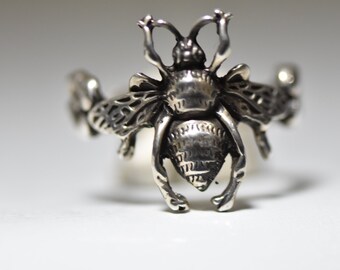 May include: A silver bee ring with intricate details. The bee is depicted in flight with its wings spread out. The ring is made of sterling silver and has a polished finish.