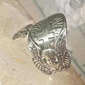 May include: A silver-toned ring featuring a cross design with sun rays, angel wings, and a cherub face. The ring has a detailed, vintage aesthetic.