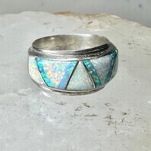 May include: A silver ring with a wide band. The ring features a mosaic design of white, blue, and green opal-like stones. The stones are arranged in a geometric pattern around the band. The ring is set on a light-colored surface.