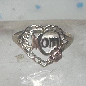 May include: A silver-toned ring featuring a heart-shaped frame with a scalloped edge. The center of the heart contains the word Mom in cutout lettering. A small, rose-colored flower accent is attached to the lower right side of the heart frame.