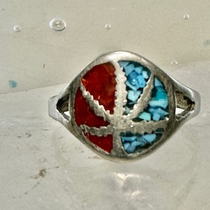 Pode incluir: Um anel de prata com uma incrustação de pedra vermelha e azul. A pedra está engastada em uma forma circular e é cercada por uma banda de prata.