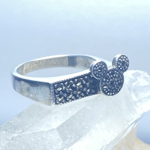 Puede incluir: Un anillo de plata con un dise&ntilde;o de cabeza de Mickey Mouse en marcasita negra. El anillo est&aacute; colocado sobre una superficie de cristal blanco.