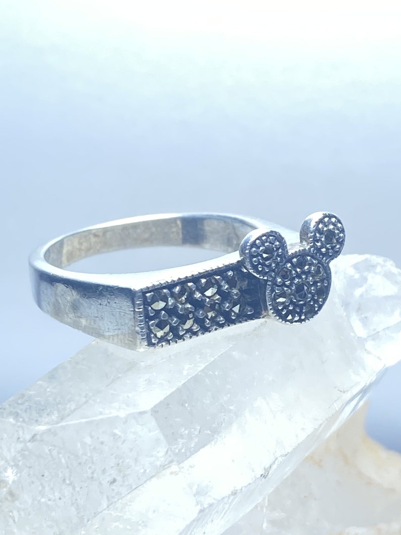 Puede incluir: Un anillo de plata con un dise&ntilde;o de cabeza de Mickey Mouse en marcasita negra. El anillo est&aacute; colocado sobre una superficie de cristal blanco.