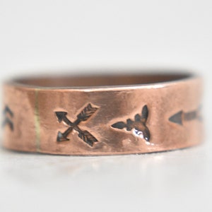 May include: A copper ring with a hammered finish and engraved arrow designs. The ring is a simple band with a slightly rounded edge.