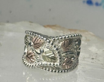 Puede incluir: Un anillo plateado con un diseño de hojas. El anillo presenta hojas alternas de color verde y oro rosa. Los bordes del anillo están bordeados con pequeñas cuentas plateadas. El anillo está sobre un fondo claro.