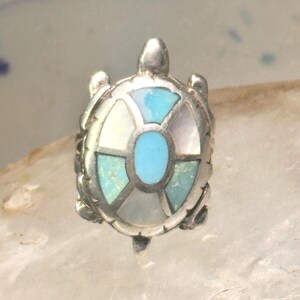 Op de afbeelding: Een zilverkleurige schildpendant met een ingelegd ontwerp van turquoise, wit en lavendel. Het schild van de schildpad heeft een segmentpatroon met een centraal ovaal van turquoise. De hanger heeft een gedetailleerde, getextureerde afwerking.