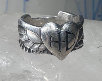 May include: A silver ring with a heart-shaped design featuring the initials "H-D" surrounded by wings.