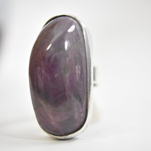 May include: A silver ring with a large, oval-shaped purple gemstone. The gemstone has a marbled pattern and a shiny finish.