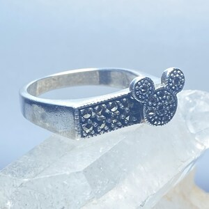 Puede incluir: Un anillo de plata con un dise&ntilde;o de Mickey Mouse en negro y plata. El anillo est&aacute; colocado sobre una superficie de cristal blanco.