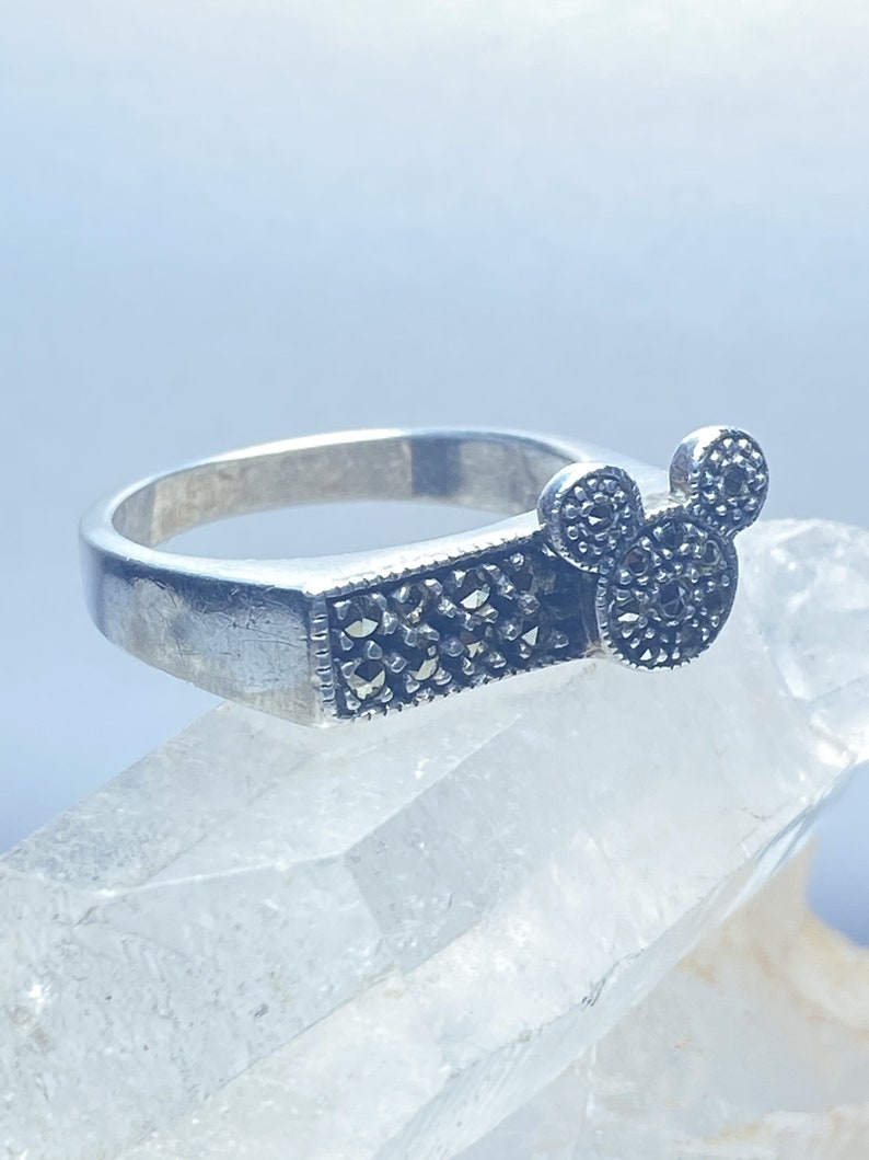 Puede incluir: Un anillo de plata con un dise&ntilde;o de Mickey Mouse en negro y plata. El anillo est&aacute; colocado sobre una superficie de cristal blanco.