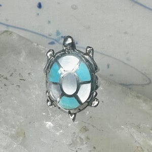 May include: A silver-toned turtle pendant with a shell design featuring turquoise, white, and gray segments. The turtle has detailed flippers, head, and tail. The pendant is likely jewelry.
