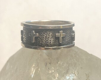May include: A silver ring with a textured band and four raised crosses. The ring is on a white background.