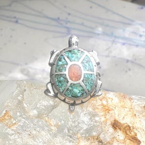May include: A silver-toned ring shaped like a turtle. The shell features a central oval orange stone surrounded by inlaid turquoise-colored stone fragments set within a segmented metal frame. The metal body displays a textured, matte finish.