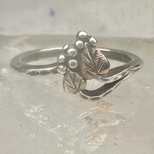 May include: A silver ring featuring a bypass band design with a hammered texture. The focal point includes two small metallic leaves in a rose gold tone, accented by a cluster of five small, smooth, round silver spheres arranged above the leaves.
