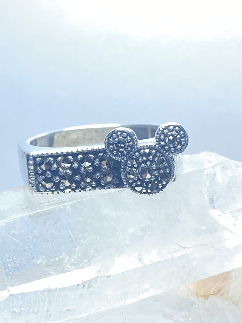Puede incluir: Un anillo de plata con un dise&ntilde;o de cabeza de Mickey Mouse en marcasita negra. El anillo est&aacute; sobre un fondo blanco.