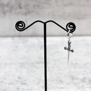 May include: A silver dagger earring hanging from a black earring stand. The earring is in the shape of a dagger with a detailed design.