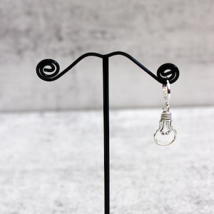 May include: A silver lightbulb charm earring hanging from a black earring stand. The stand has a decorative base.