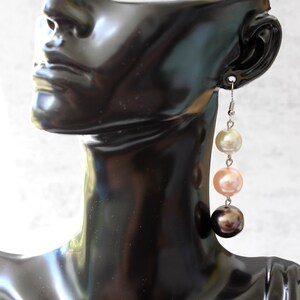 May include: A dangling earring featuring four graduated pearls in cream, peach, and dark brown hues. The earring is suspended from a silver-colored hook and displayed on a black mannequin head.