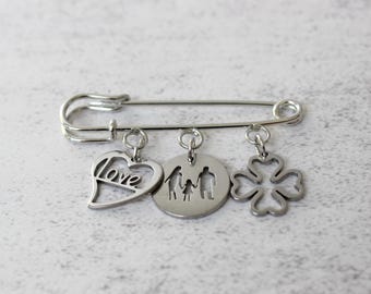 Pode incluir: Alfinete de segurança prateado com três pingentes pendurados. Os pingentes incluem um coração com a palavra "love", um pingente circular com uma silhueta familiar e um trevo de quatro folhas. Em uma superfície texturizada cinza claro.
