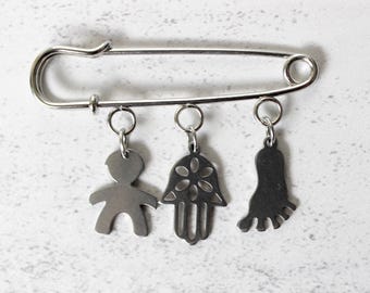 Pode incluir: Um alfinete de segurança prateado com três pingentes pendurados: uma figura de palito, uma mão de Hamsa e um pé. Os pingentes são suspensos por pequenos anéis presos ao alfinete.