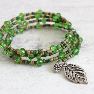 Puede incluir: Un brazalete de envoltura de cuentas verdes con un dije de hoja de plata. El brazalete está hecho con múltiples hilos de cuentas verdes y marrones.