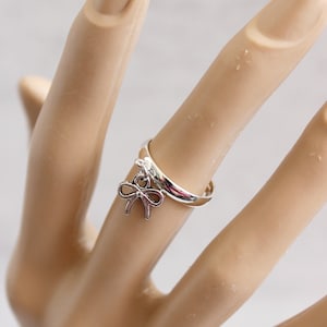 May include: A silver ring with a bow charm. The ring is on a finger, and the bow charm hangs from the ring. The ring is a simple band with a small, dangling bow. The bow is a delicate, open design.