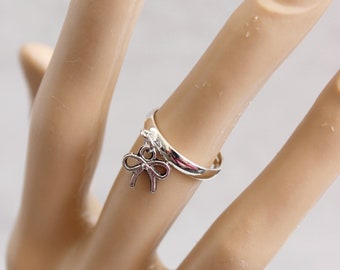 May include: A silver ring featuring a bow pendant. The ring is worn on a finger, with the bow charm suspended from the band. The bow is a delicate, open design. The ring is a simple band. The bow charm adds a touch of elegance.