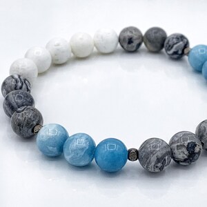 May include: A white, gray, and blue gemstone bracelet with a gradient of color. The bracelet is made of round beads and has a simple design.