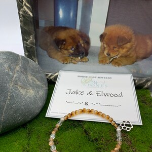 May include: A brown and clear beaded bracelet with a silver paw print charm. The bracelet is on a green surface with a card that says "Morse Code Jewelry Cedar Stones Jake & Elwood" and a picture of two puppies in a frame.