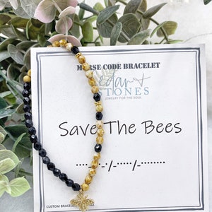 May include: A black and gold beaded bracelet with a bee charm. The bracelet is on a white card with the text "Morse Code Bracelet", "Cedar Stones", "Jewelry for the Soul", "Save The Bees", and "... -/--/--..." in black text.