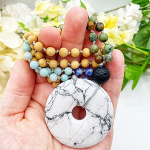 May include: A white howlite donut pendant with a hole in the center, hanging from a multi-colored beaded necklace. The necklace features wooden beads, blue lapis lazuli beads, green turquoise beads, and brown jasper beads.