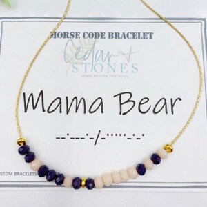 May include: A gold chain bracelet with purple and white beads. The bracelet is on a white card with the text "Morse Code Bracelet", "Cedar+ Stones", "Jewelry for the Soul", "Mama Bear", and "..------/---.." in black.