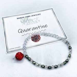 May include: A clear glass beaded bracelet with a red rhinestone charm. The bracelet is on a white card with the text "Morse Code Jewelry Cedar Stones Jewelry for the Soul Quarantine" and a dashed line.