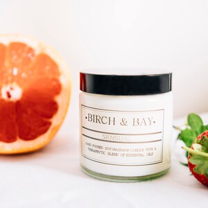 May include: A white soy massage candle in a glass jar with a black lid. The candle is labeled "Birch & Bay" and "Sensual" with the text "Hand-poured soy massage candle with a therapeutic blend of essential oils".