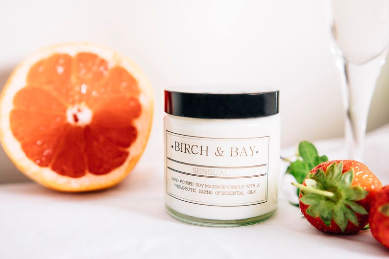May include: A white soy massage candle in a glass jar with a black lid. The candle is labeled "Birch & Bay" and "Sensual" with the text "Hand-poured soy massage candle with a therapeutic blend of essential oils".