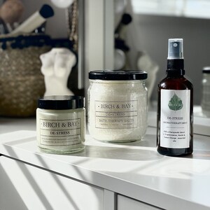 May include: Three glass jars with black lids and white labels. The labels read "Birch & Bay" and "De-Stress". The jars contain bath salts, a soy massage candle, and an aromatherapy mist.