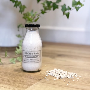 May include: A glass bottle with a black lid filled with a white, creamy bath soak. The label reads "Birch & Bay - Oat & Coconut Milk Bath - Natural // Vegan // Handmade - Perfect for dry, itchy & eczema prone skin". A small pile of oats is in the foreground.