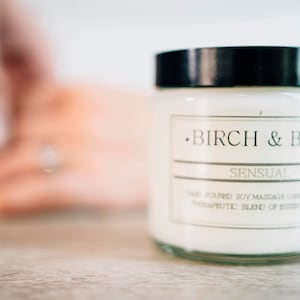 May include: A white candle jar with a black lid and a label that reads "Birch & Bay" and "Sensual" Hand Poured Soy Massage Candle with a Therapeutic Blend of Essential Oils.