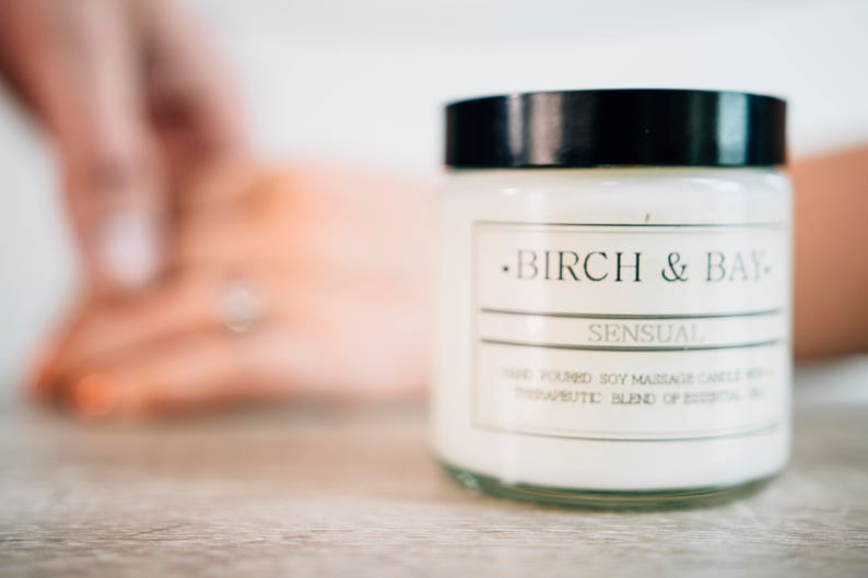 May include: A white candle jar with a black lid and a label that reads "Birch & Bay" and "Sensual" Hand Poured Soy Massage Candle with a Therapeutic Blend of Essential Oils.