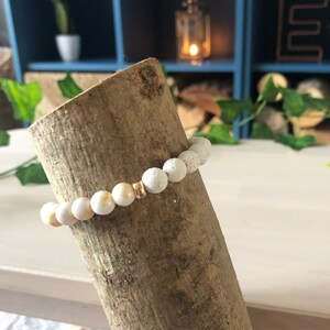 May include: White lava rock beaded bracelet with a gold accent bead. The bracelet is displayed on a wooden log.