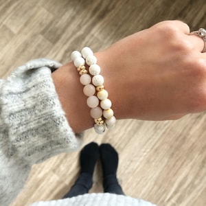 May include: Two bracelets stacked on a wrist. The top bracelet is made of white lava beads with gold spacers. The bottom bracelet is made of light pink and white beads with gold spacers.