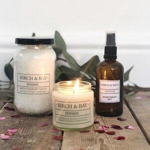 May include: Aromatherapy set featuring a lit candle, bath salts, and a mist spray bottle. The candle and spray bottle are amber-colored glass. The bath salts are in a clear jar. All items have labels that read "Birch & Bay Sensual".