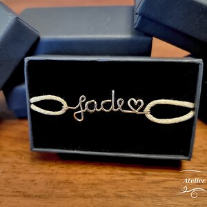 May include: A gold wire bracelet with the name "Jade" spelled out and a heart charm. The bracelet is on a white background in a black box.
