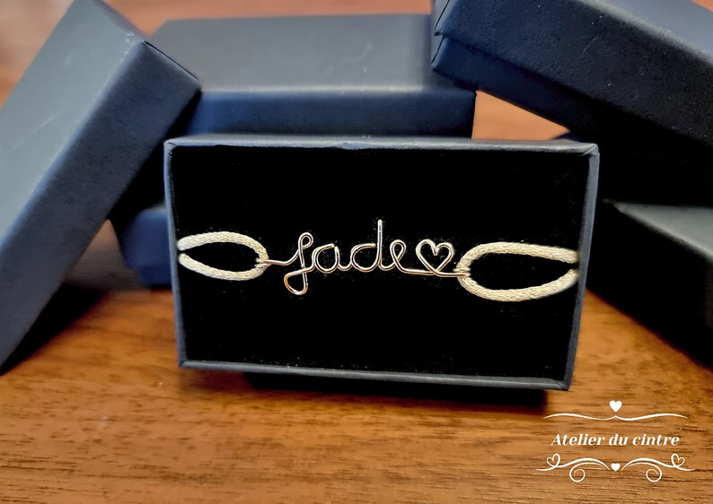 May include: A gold wire bracelet with the name "Jade" spelled out and a heart charm. The bracelet is on a white background in a black box.