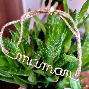 May include: A gold-toned bracelet with a delicate chain and the word "maman" spelled out in wire. The bracelet is resting on a green plant.