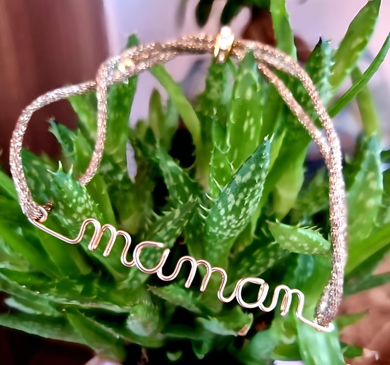 May include: A gold-toned bracelet with a delicate chain and the word "maman" spelled out in wire. The bracelet is resting on a green plant.