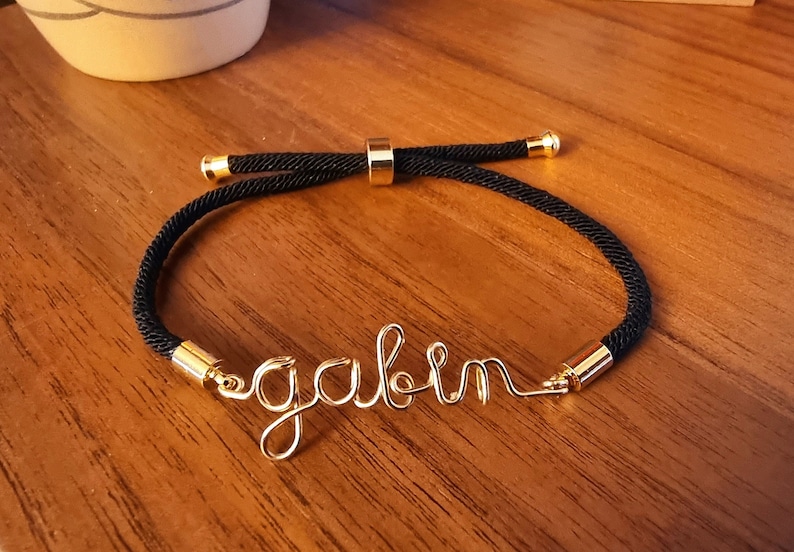 Puede incluir: Una pulsera de cuerda negra con detalles dorados y el nombre "gabin" en alambre dorado. La pulsera es ajustable y descansa sobre una superficie de madera.