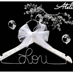 May include: White wooden hanger with a white tulle bow and rhinestones. The hanger has the word "Lou" written in silver wire and the date "10.10.2016" engraved on the bottom.