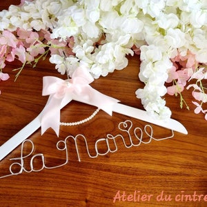 May include: White wooden hanger with a pink ribbon and a wire inscription that says "La Marie" with a heart at the end. The hanger is decorated with a pearl necklace. The hanger is on a wooden surface with pink and white flowers.